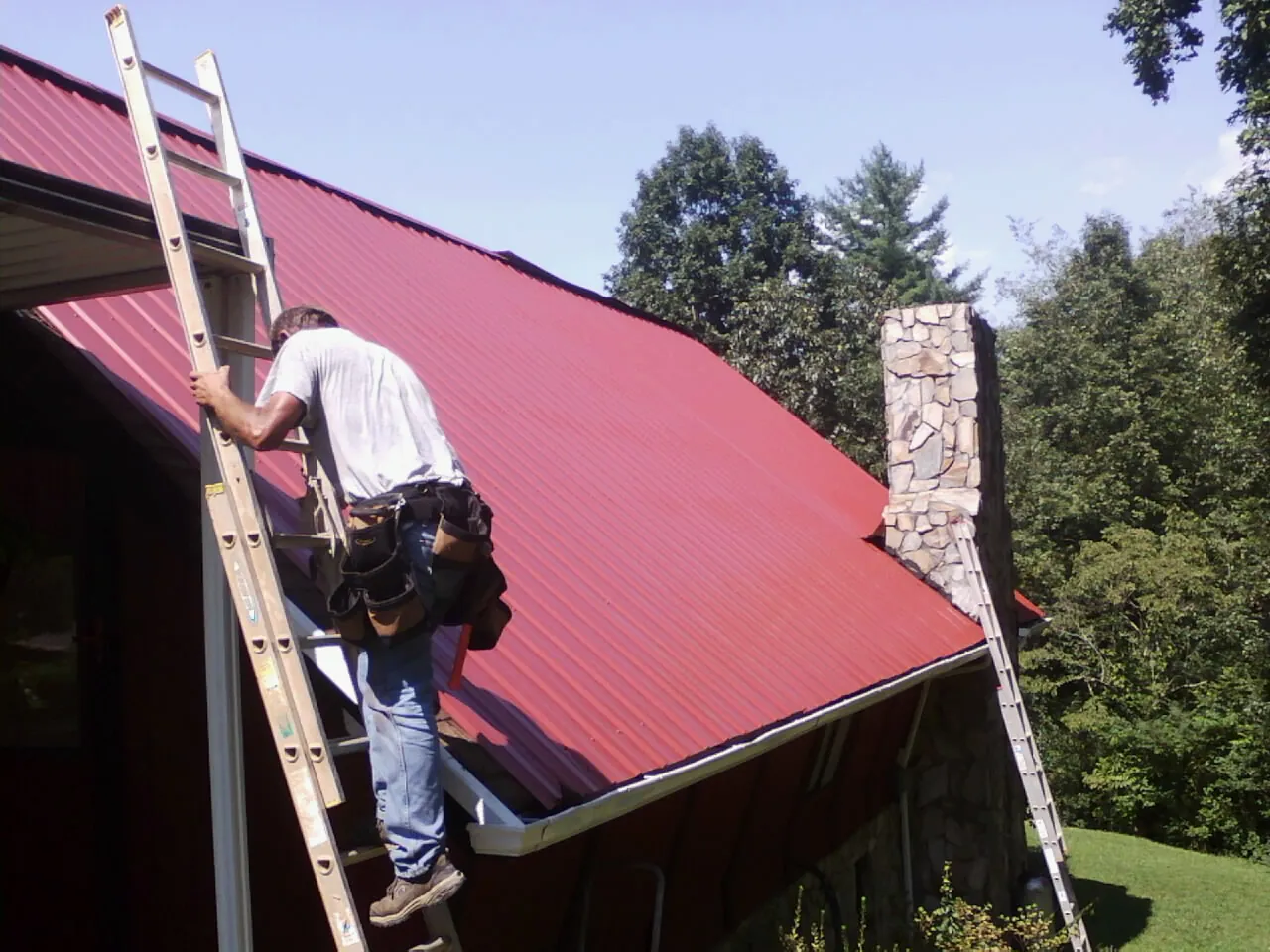 Roofer on a ladder beside a red metal roof and stone chimney — Soffit & Fascia Repair by Roofing Brothers in Akron